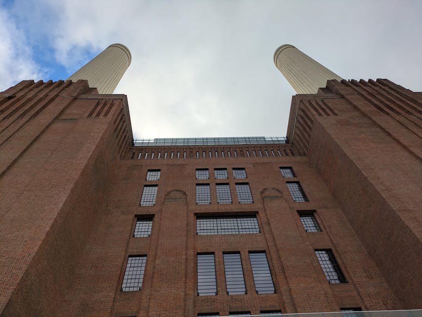 A large historic brick building with a distinctive industrial design, featuring four towering white smokestacks rising above the structure. The building has a range of rectangular windows with dark fr